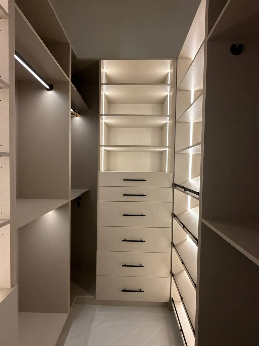 Beige walk-in closet with a central drawer tower, lit shelves and hanging sections on both sides