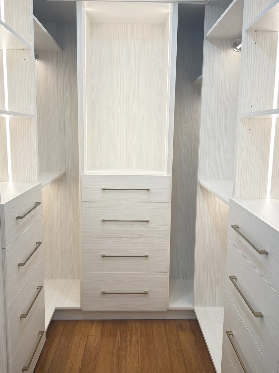 White walk-in closet with lit shelving on both sides, a drawer tower at the end wall and an oak floor